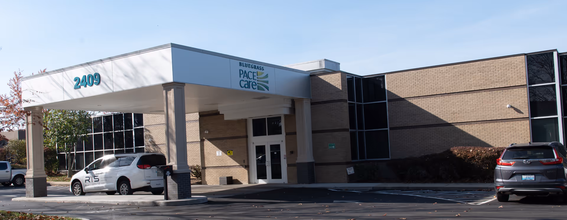 Exterior view of Bluegrass PACE Care entrance with canopy and parked cars.