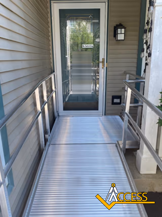 Metal ramp with handrails leads to glass door entry on beige building exterior.