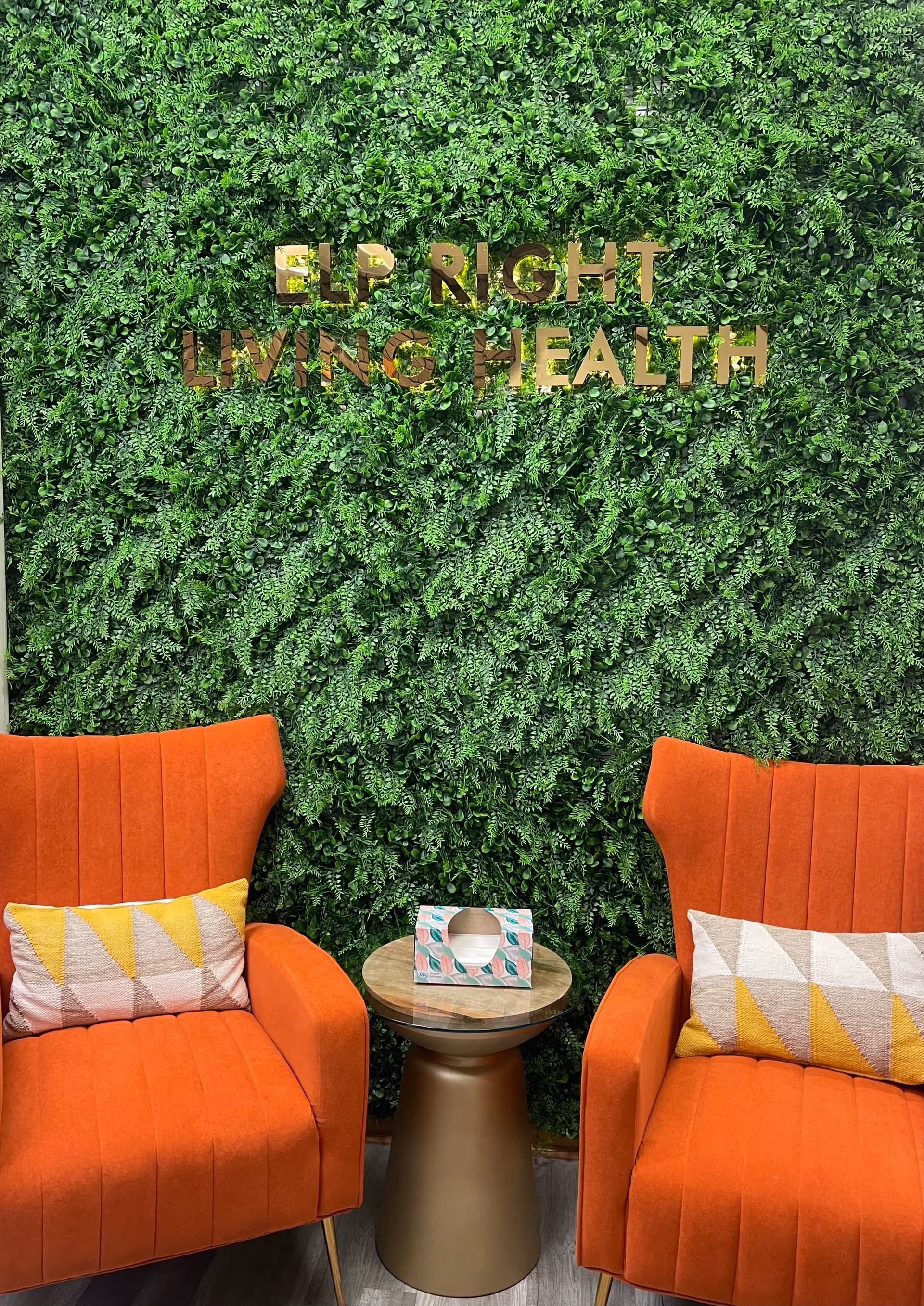 Two orange chairs flank a gold side table against a green living wall with gold sign.