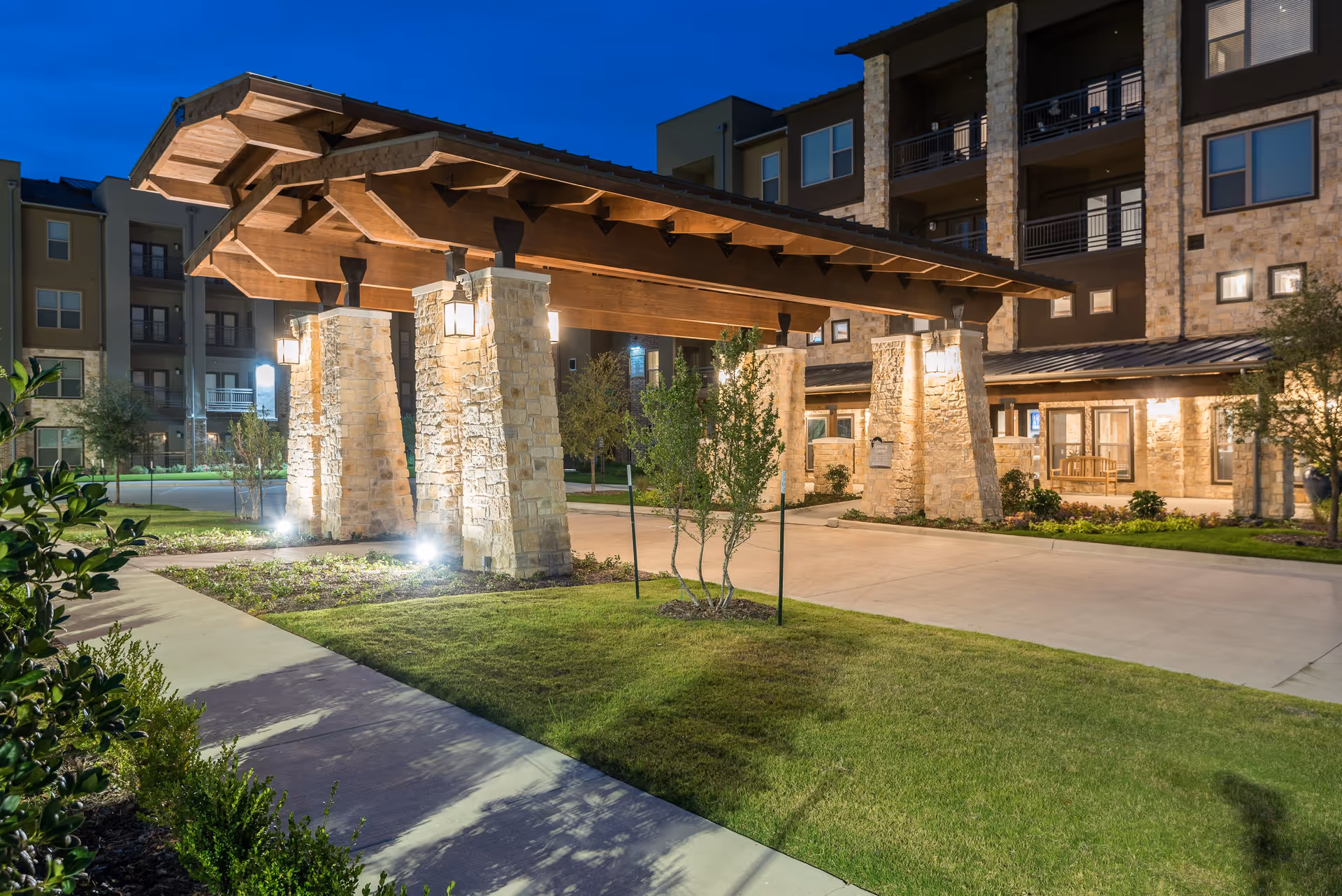 Stone-pillared covered porte-cochere and landscaped driveway at the front of a multi-story senior living building lit at dusk.
