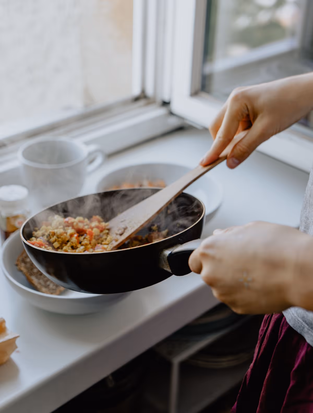 Person cooks vegetables in a pan on a kitchen stove by a window.