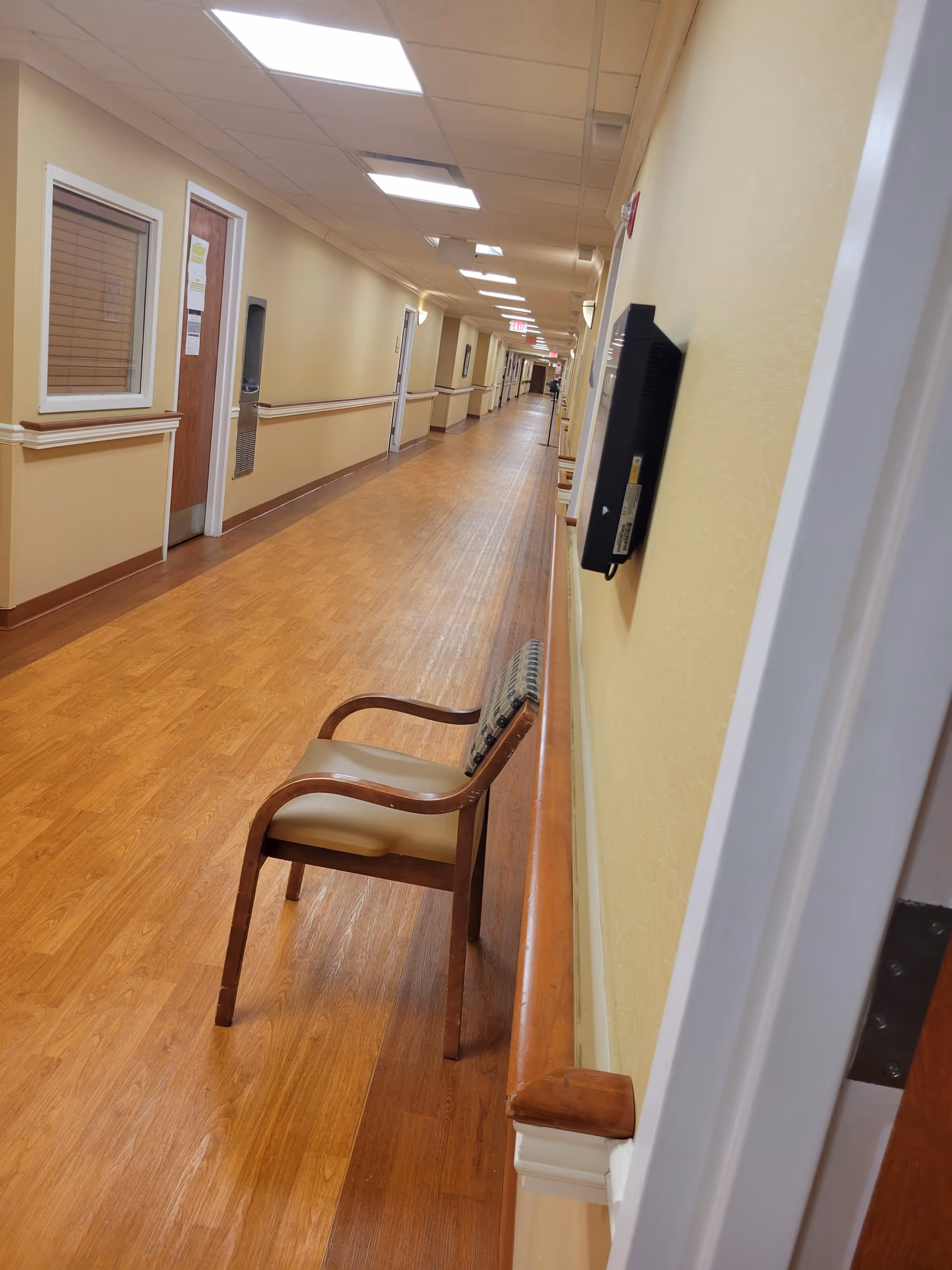 A long, empty hallway in a senior care facility with a wooden chair.