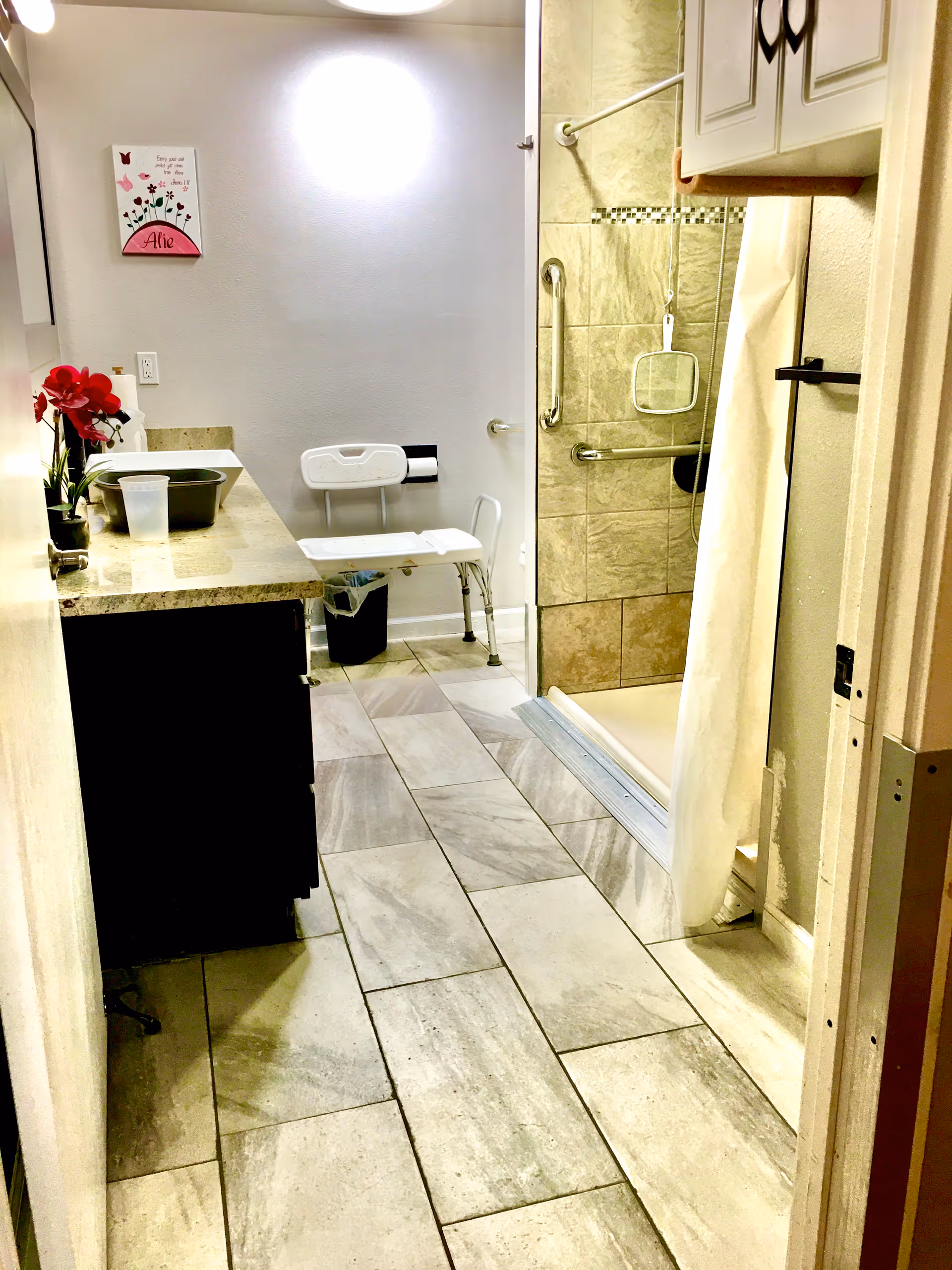 Bright bathroom in senior living facility with sink, vanity, shower, grab bars, and chair.
