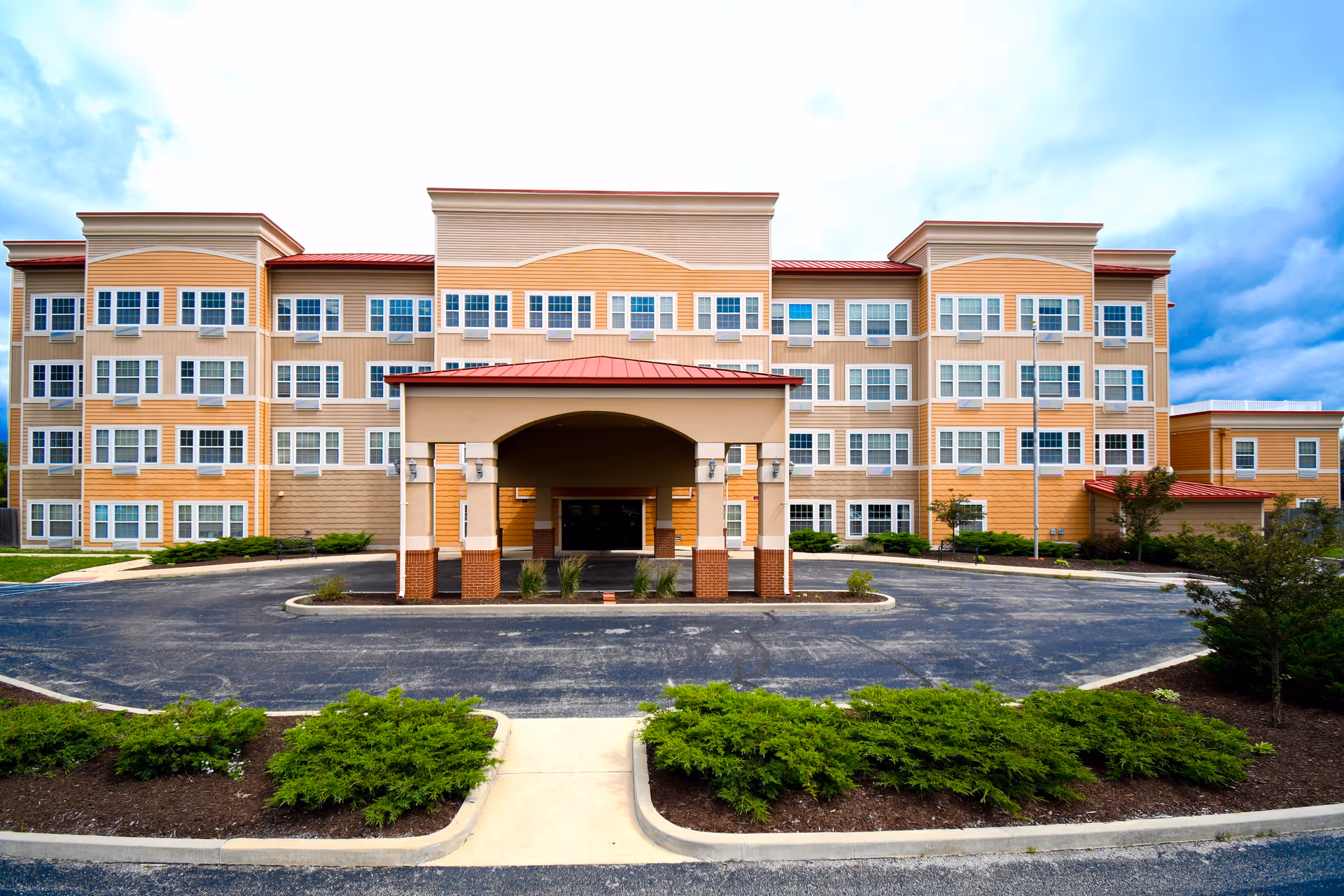 Four-story beige and tan senior living building with a covered porte-cochère entrance and circular driveway.