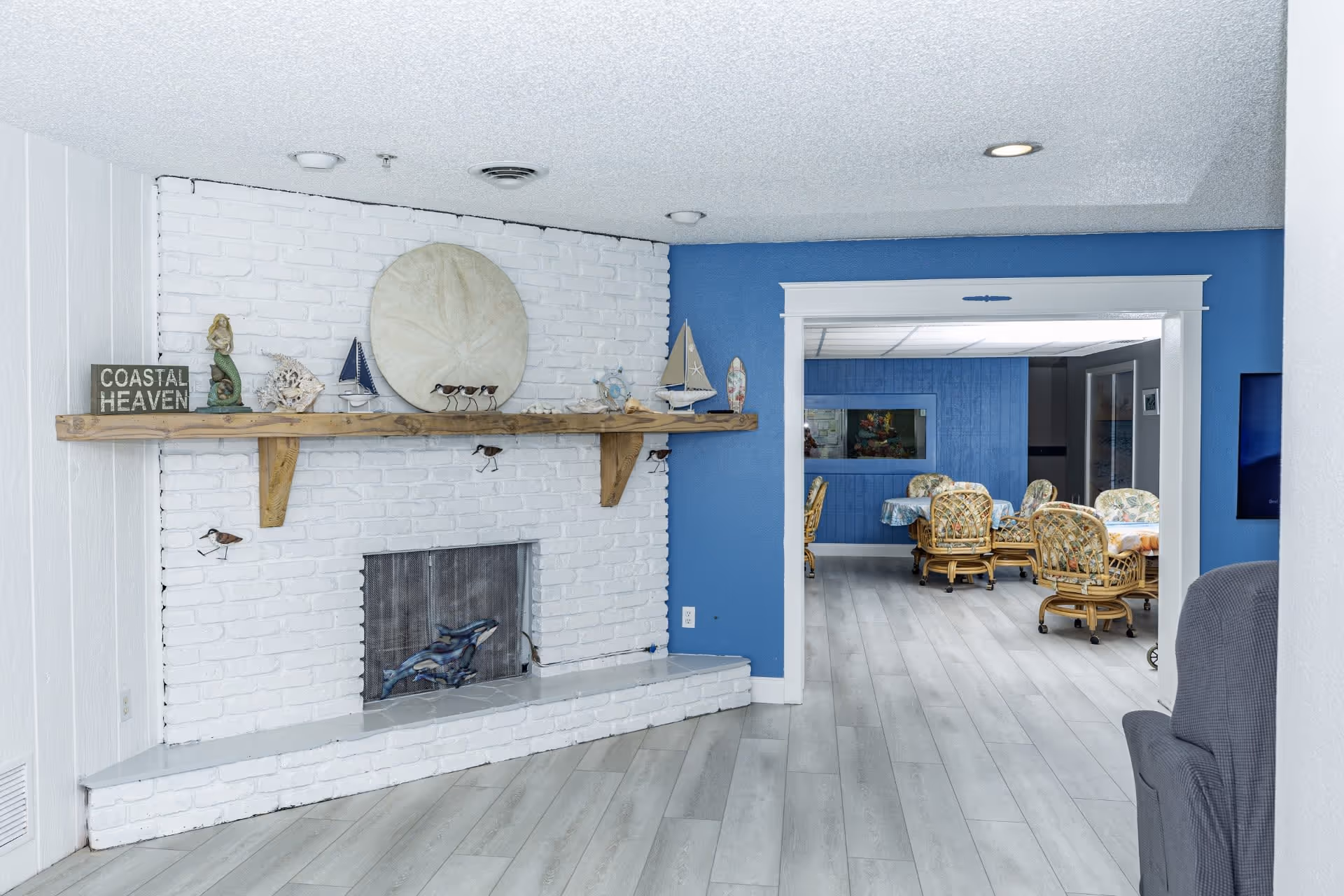 Interior view of a senior living facility showing a white brick fireplace with a wooden mantel decorated with coastal-themed items including a mermaid figurine, a sand dollar, and small sailboats. The room has light gray wood flooring and blue walls. Through an open doorway, a dining area with tables covered in floral tablecloths and wicker chairs with floral cushions is visible.