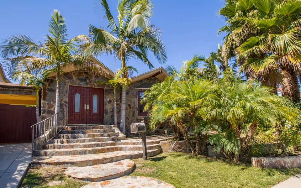 Stone-faced building entrance with red double doors, curved stone steps, a mailbox and palm trees in the front yard.