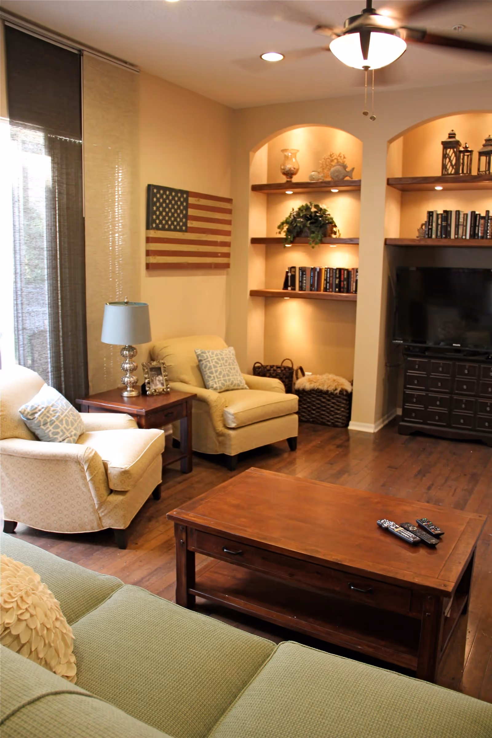 Cozy living room with upholstered chairs and sofa, a wooden coffee table, built-in lit shelves, and a TV.