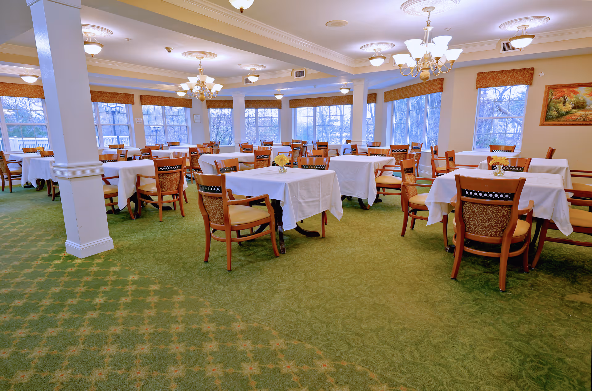 A spacious dining room with multiple tables covered in white tablecloths, each surrounded by wooden chairs with cushioned seats. The room has large windows letting in natural light, green patterned carpet, and several chandeliers hanging from the ceiling. A painting is visible on one wall.