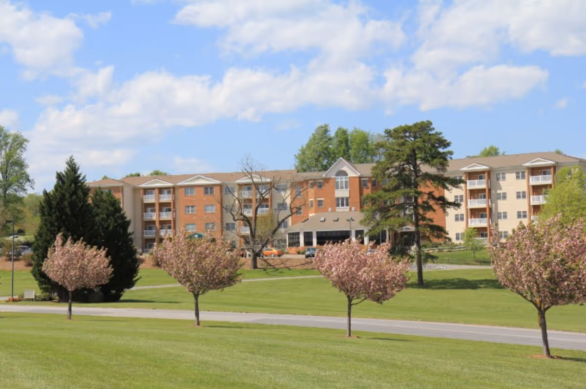 Exterior view of King's Grant Retirement Community building with a well-maintained lawn and several blossoming trees in the foreground under a partly cloudy blue sky.