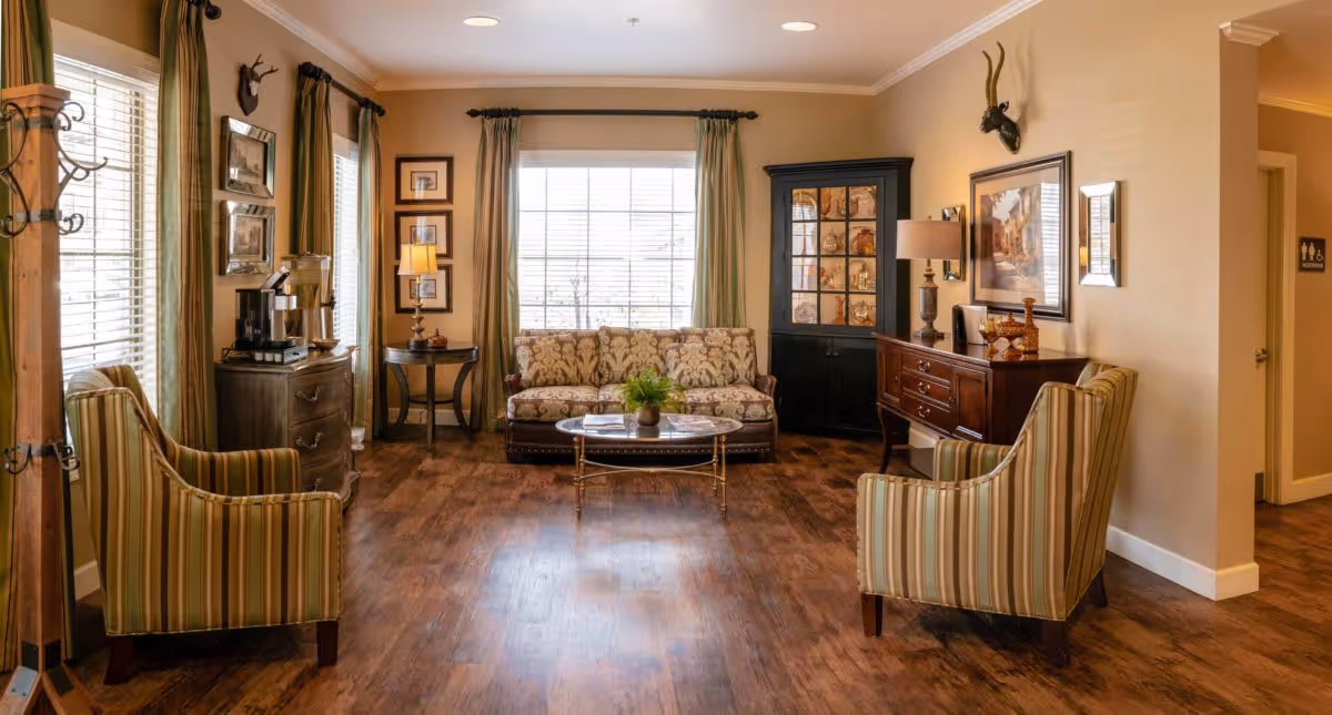 A cozy living room area with wooden flooring, two striped armchairs facing a floral-patterned sofa. There is a glass coffee table with a small plant in the center. The room has large windows with green curtains, a wooden cabinet with glass doors, a side table with a lamp, and framed artwork on the walls.