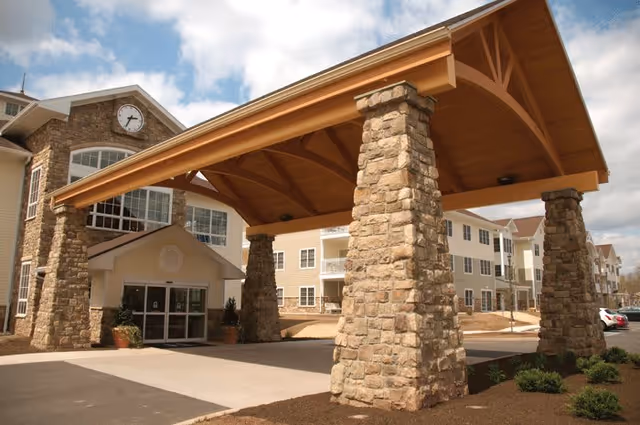 Entrance of The Heritage of Green Hills facility featuring a large covered driveway supported by stone pillars, with a multi-story building in the background under a partly cloudy sky.