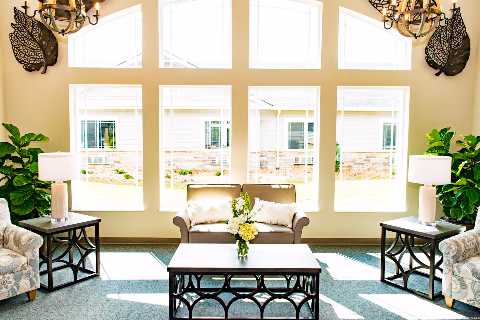 A bright and inviting living room area with large windows letting in natural light. The room features a small sofa with two pillows, a coffee table with a vase of flowers, two side tables each with a lamp, two patterned armchairs, and green plants on either side. Decorative wall sconces shaped like leaves are mounted on the walls.