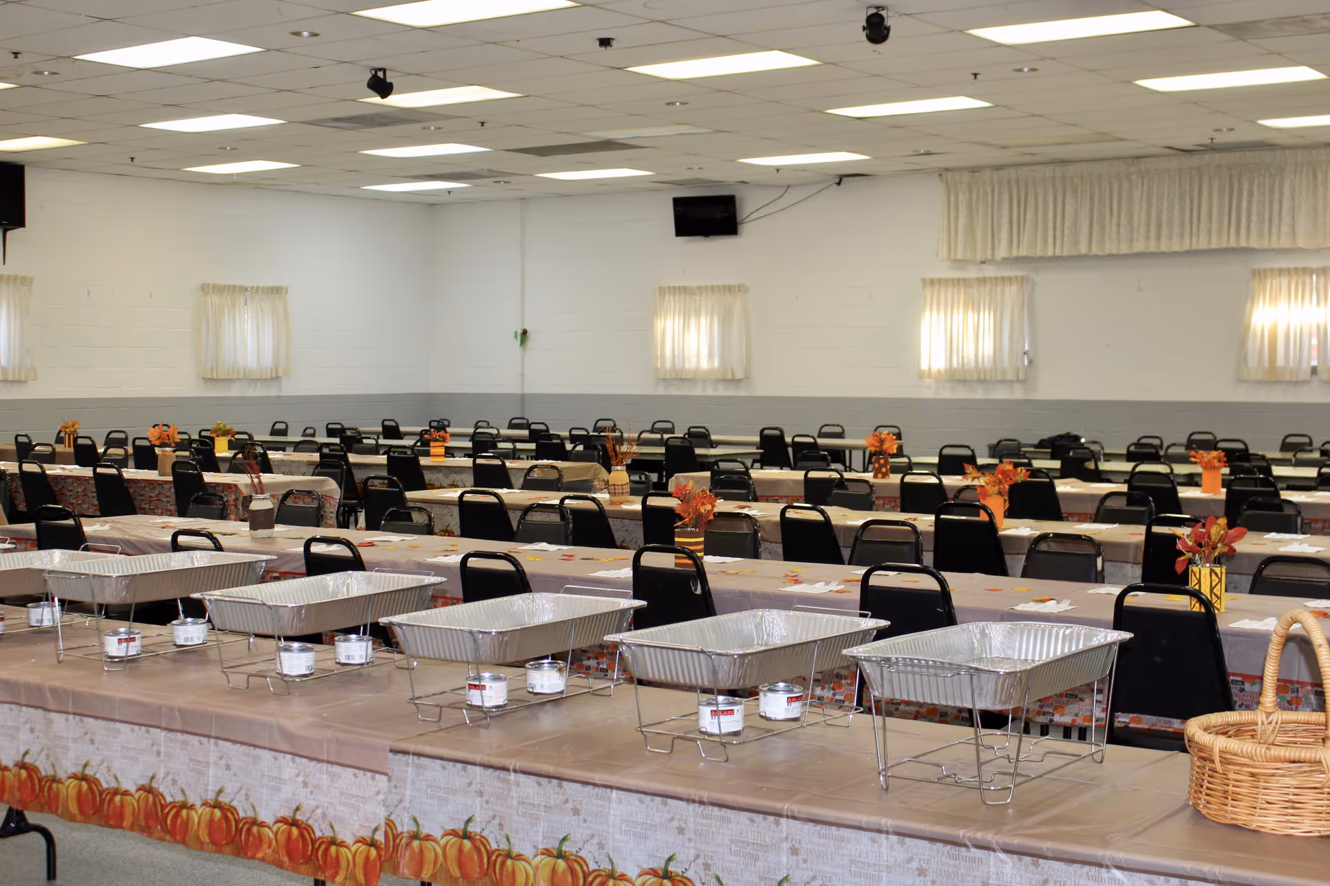 A large indoor dining hall with long banquet tables, chafing dishes, and autumn decor.