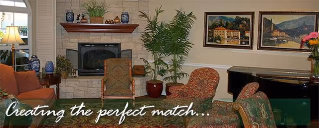 Cozy living room in a senior facility featuring a fireplace, plants, chairs, and artwork.
