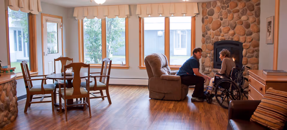 A cozy senior living room with a stone fireplace, dining table, and residents chatting.