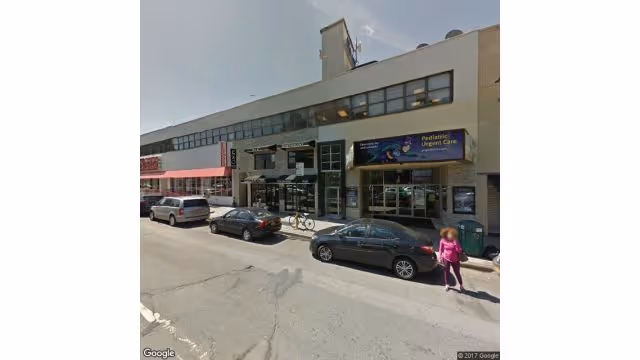 Exterior view of a storefront block with cars parked and a pediatric urgent care.
