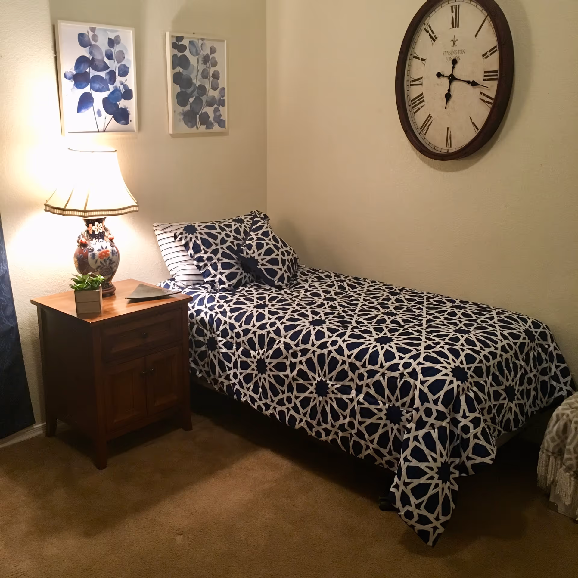 A cozy bedroom with a single bed covered in a blue and white patterned bedspread and matching pillows. Next to the bed is a wooden nightstand with a decorative lamp and a small potted plant. Two framed blue botanical prints hang on the wall above the nightstand, and a large round wall clock with Roman numerals is mounted on the wall above the bed.