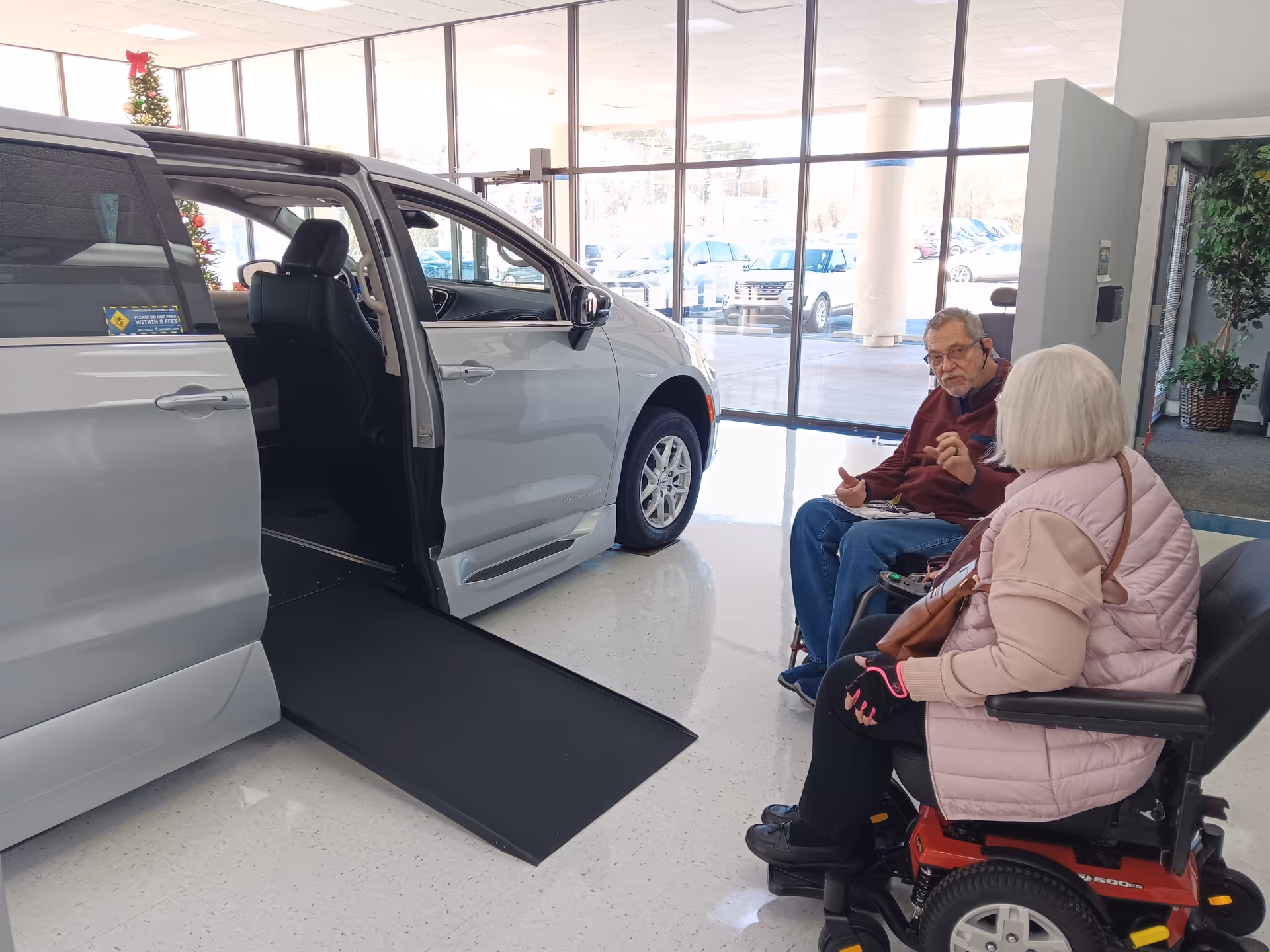 Inside a showroom, a white wheelchair-accessible vehicle with a ramp, as two seniors converse.