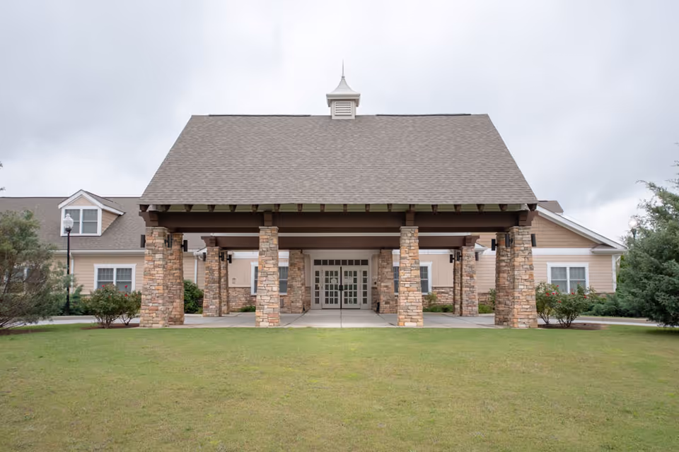 Front exterior view of a building with a large covered entrance supported by stone pillars, surrounded by green grass and shrubs under a cloudy sky.