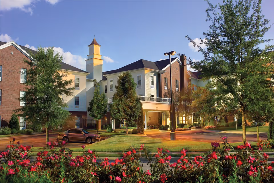 Exterior view of Belmont Village Senior Living St. Matthews building with a mix of brick and light-colored siding, surrounded by trees, flowers, and a parked car under a clear blue sky.