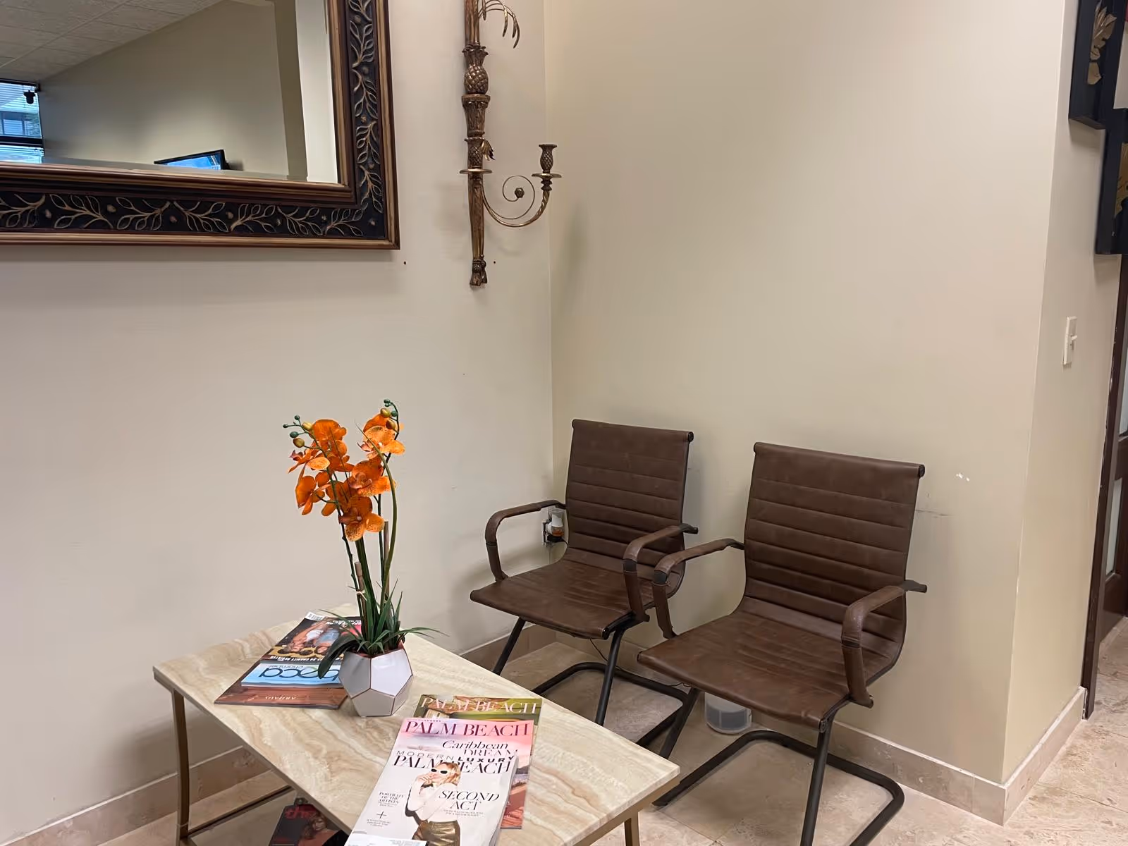 Two brown waiting chairs beside a coffee table with magazines and orange flowers.