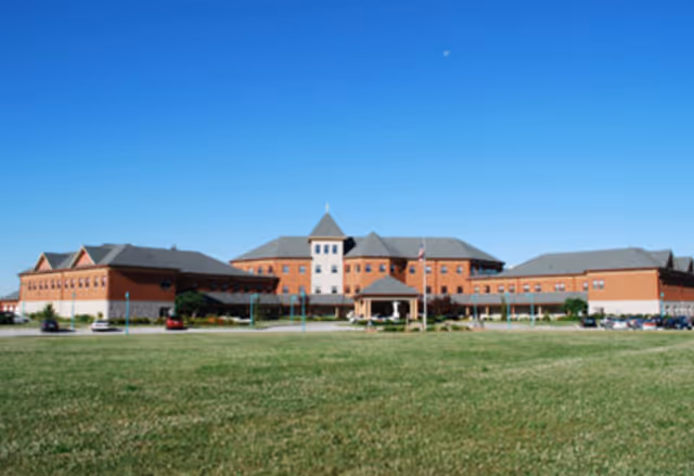 Front view of a large brick senior living facility with a central entrance, drive, and expansive lawn under a clear blue sky.