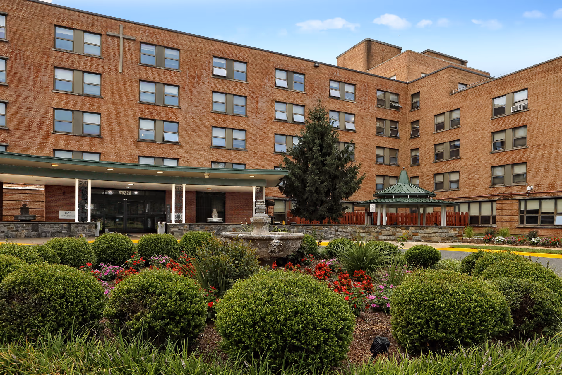 Exterior view of a multi-story brick building with many windows, a covered entrance, and a landscaped garden with trimmed bushes, flowers, and a stone fountain in front.