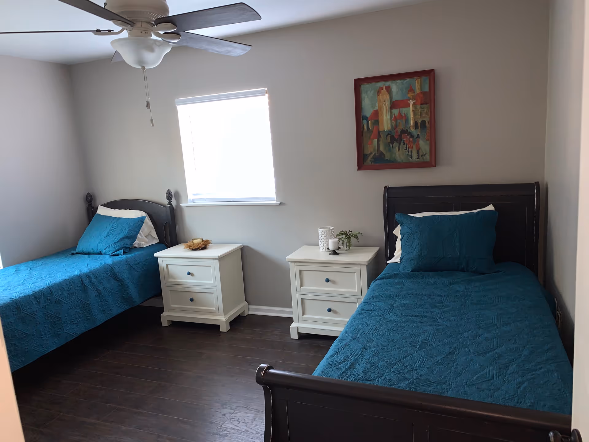 A bedroom with two single beds, each with dark wooden frames and teal bedspreads. Between the beds are two white nightstands with drawers, one holding a candle and a small plant. A window with blinds is centered on the wall above the nightstands, and a colorful framed painting hangs on the wall above the right bed. A ceiling fan with a light fixture is visible on the ceiling.