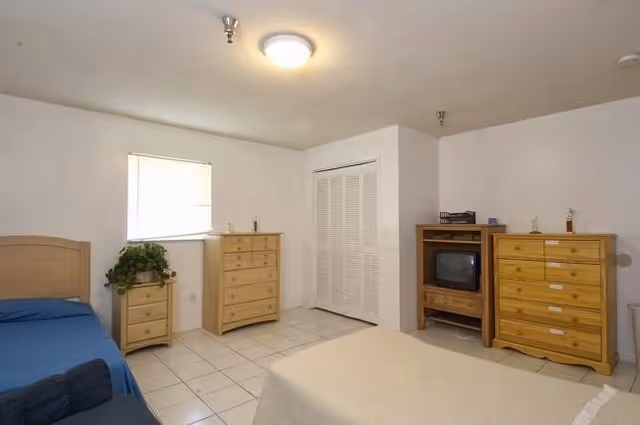 A simple, sparsely furnished bedroom with two dressers, a bed, plant, and a small TV.
