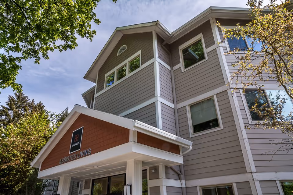 Exterior view of Marquis Wilsonville Assisted Living facility showing a multi-story building with gray siding, white trim, and a covered entrance with a sign that reads 'ASSISTED LIVING'. Trees with green leaves surround the building under a partly cloudy sky.