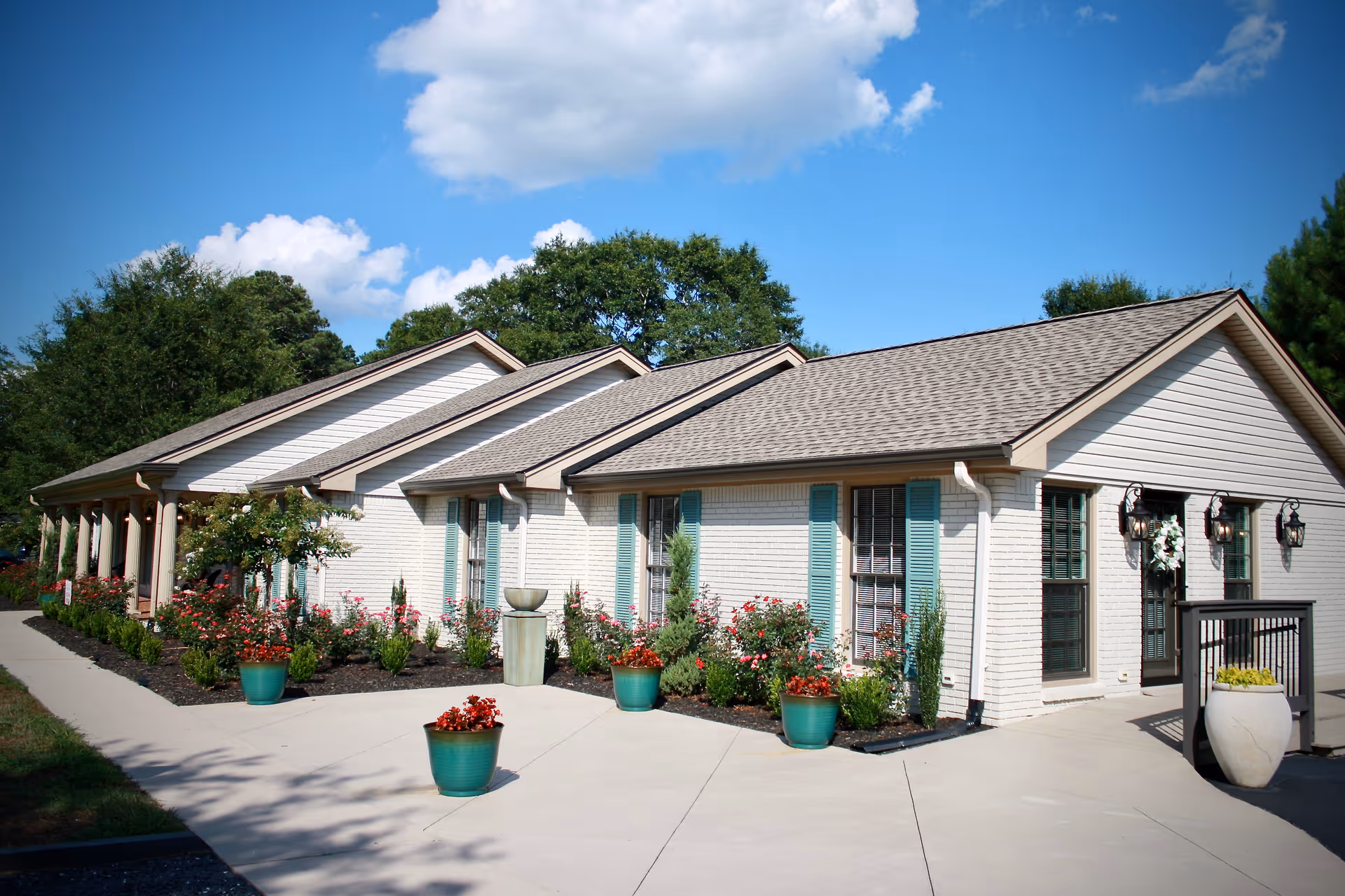 Exterior view of a single-story senior living facility building with white brick walls, teal window shutters, and a gray shingled roof. The building is surrounded by a well-maintained garden with various flowering plants and potted flowers along a concrete walkway. The sky is clear with a few clouds, and trees are visible in the background.