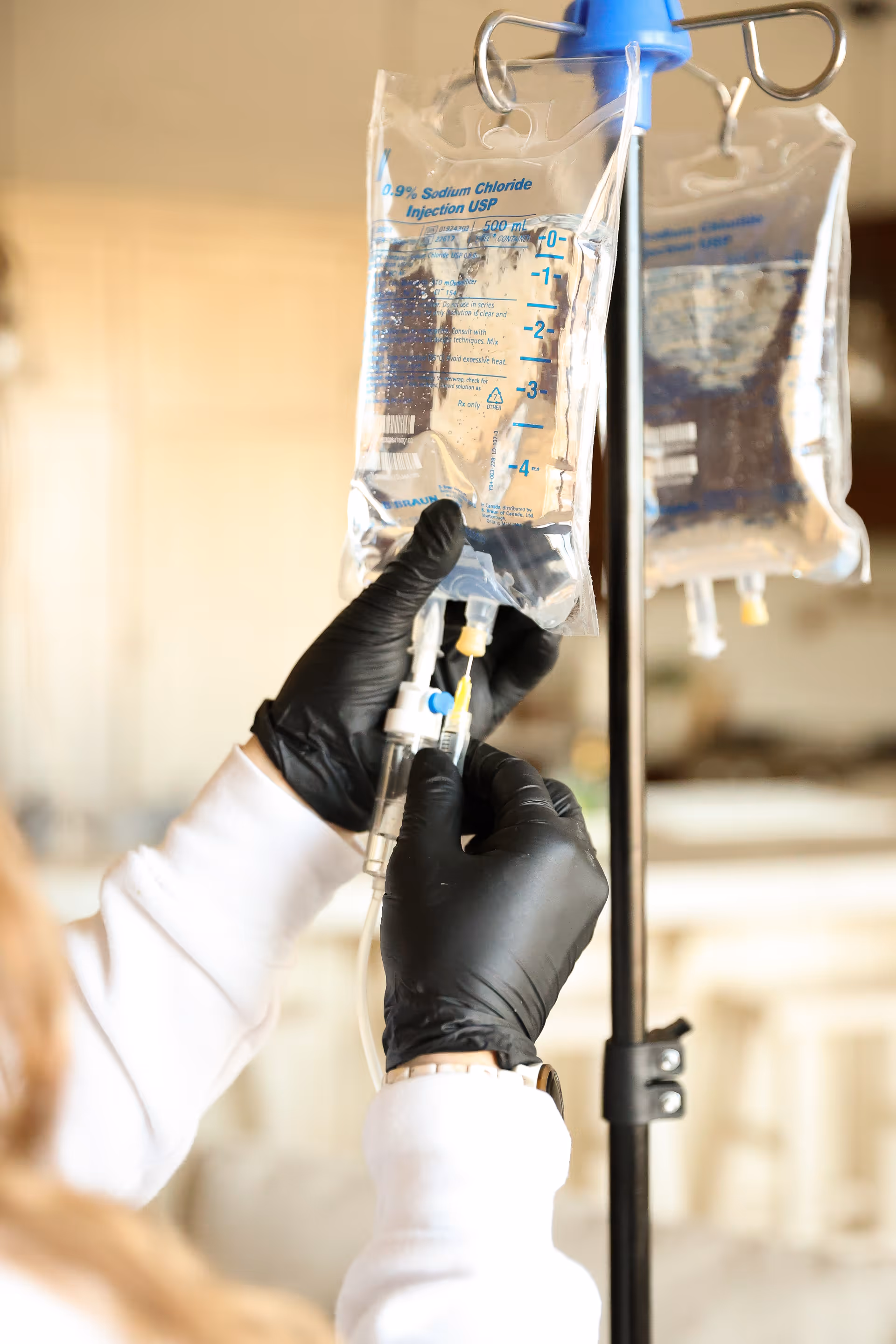 A clinician in gloves adjusts an IV bag on a pole in a hospital room.