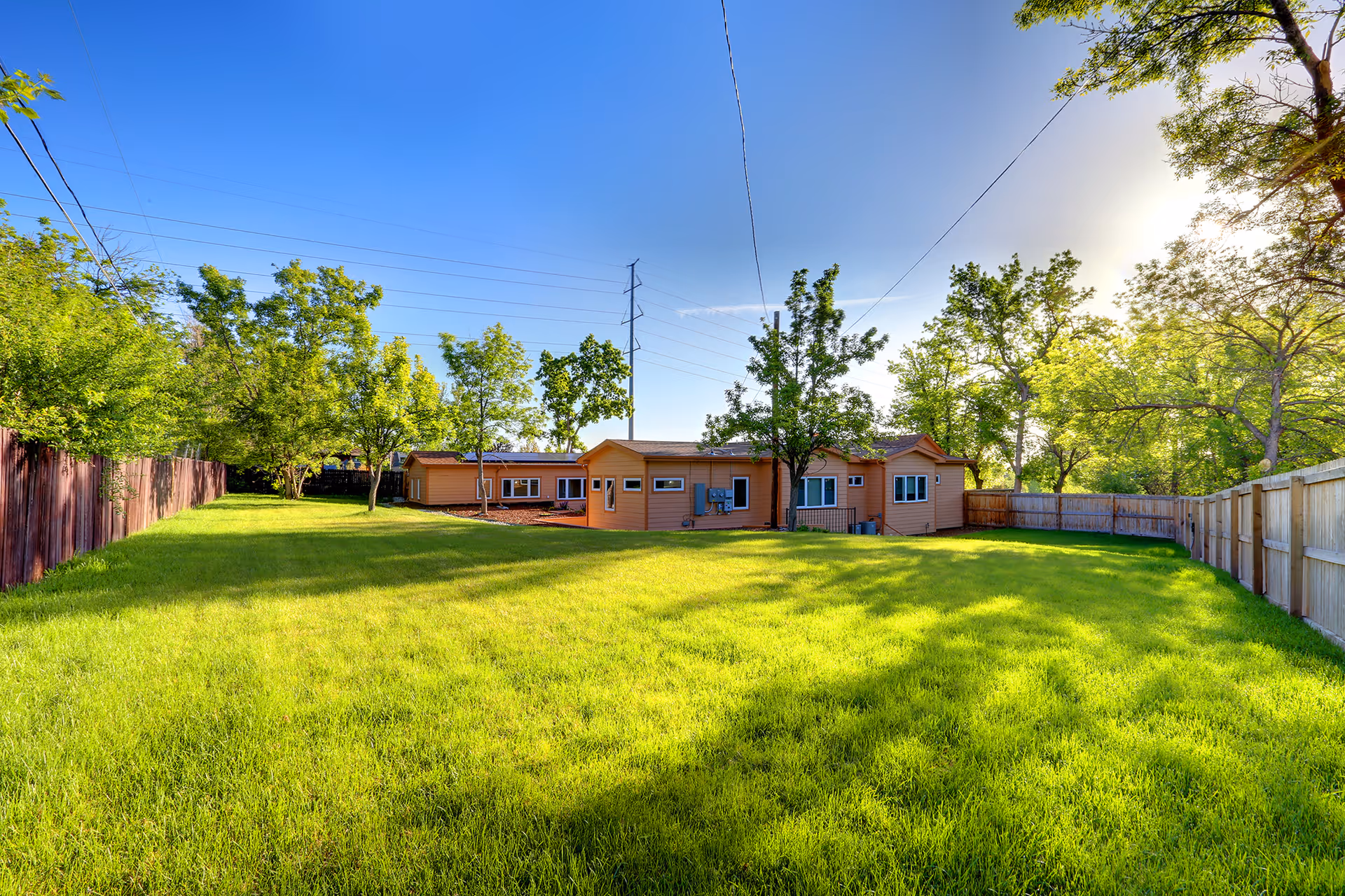 A large, well-maintained grassy backyard area with several trees and a wooden fence surrounding the space. In the background, there is a single-story building with a light brown exterior under a clear blue sky.
