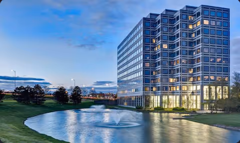 Modern glass-fronted senior living building near a pond with a fountain.