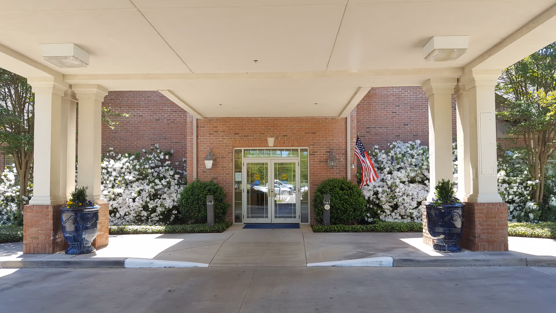 Entrance to a brick building with columns, flowering shrubs, and an American flag.