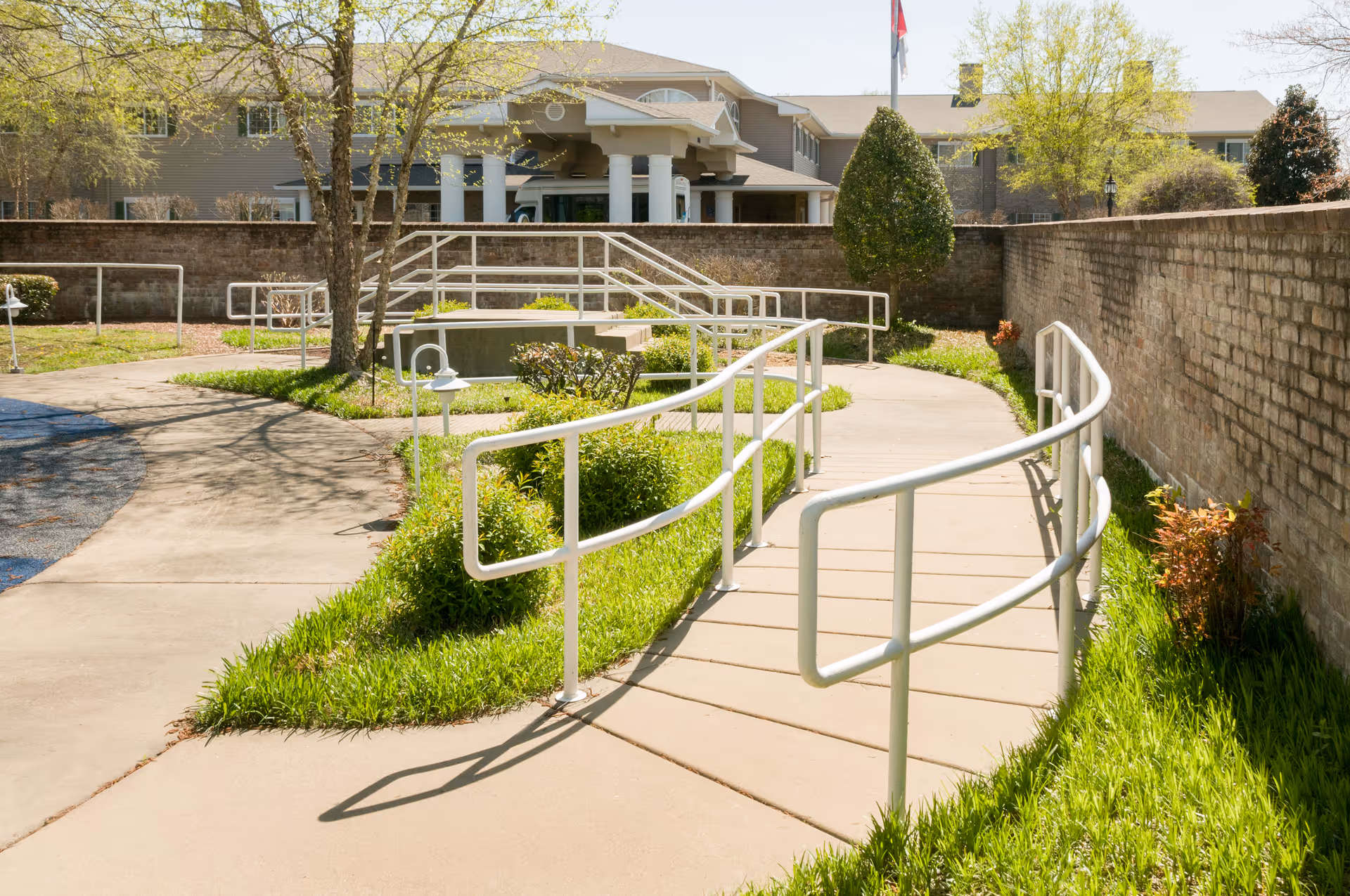 Curved concrete walkway with white handrails and landscaping leading to the entrance of a senior living facility.