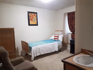 Single-occupancy assisted living bedroom with a twin bed, recliner, dresser, sink, and framed wall art.