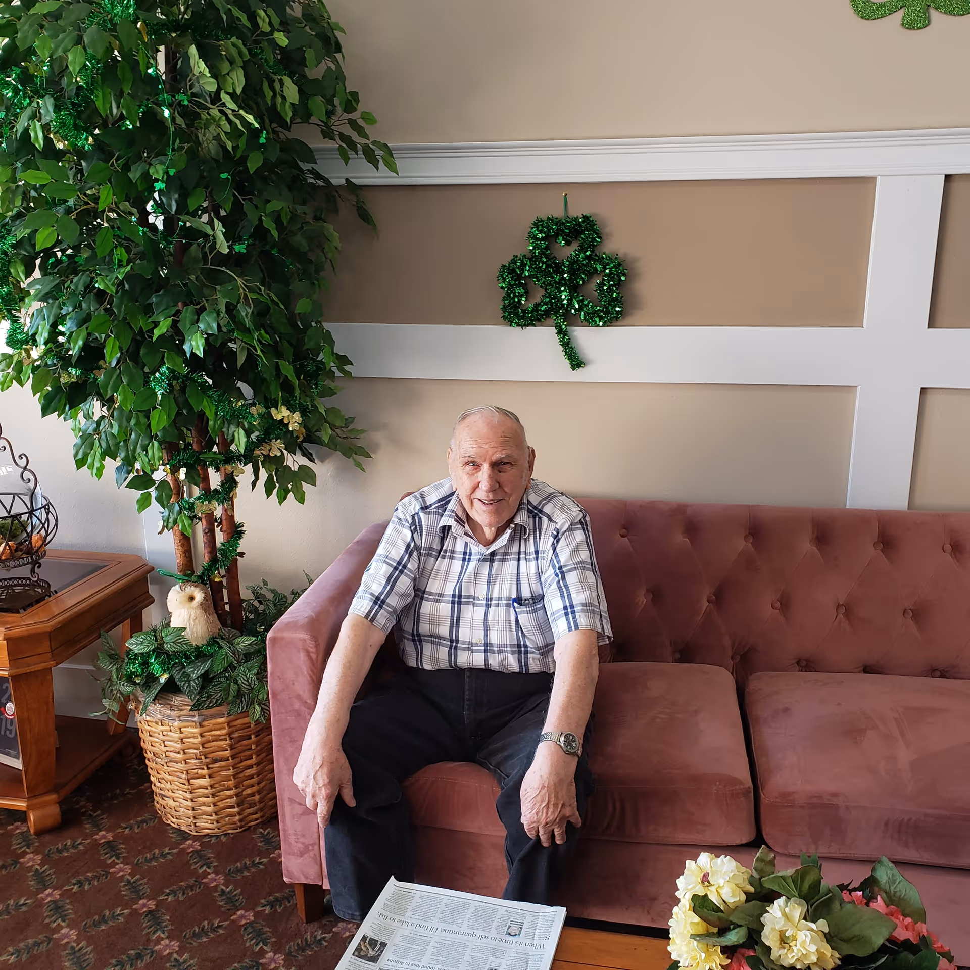 An elderly man sits on a pink tufted sofa in a decorated common area with plants and a shamrock on the wall.