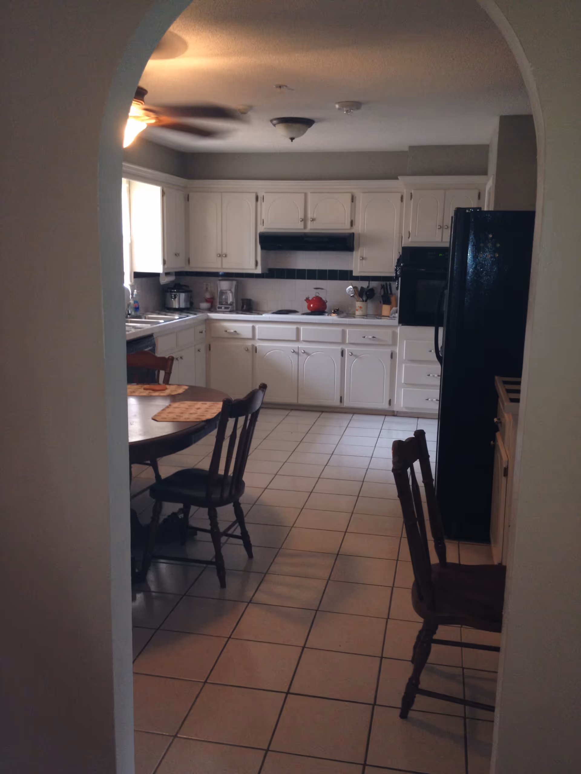 View through an arched doorway into a kitchen with white cabinets, a black refrigerator, a stove with a red kettle, and a round wooden dining table with chairs. The floor is tiled and there is a ceiling fan with a light.