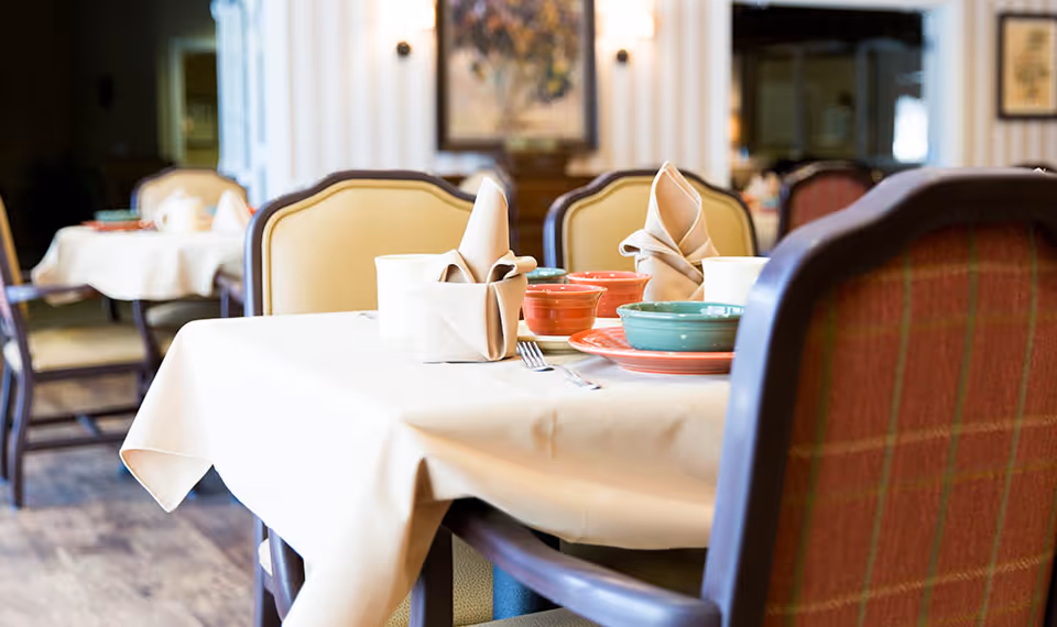 A dining room table set with beige tablecloth, folded beige napkins, colorful bowls and plates, and silverware. The chairs around the table have cushioned backs with a plaid pattern. Additional tables and chairs are visible in the softly lit background.