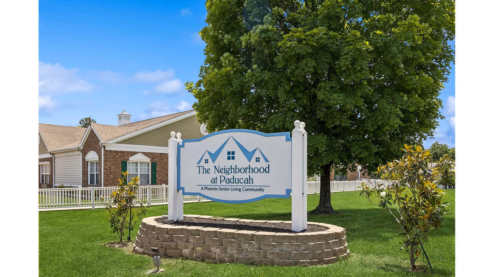 A white and blue entrance sign reading "The Neighborhood at Paducah" on a raised stone bed with a large tree and senior living buildings in the background.