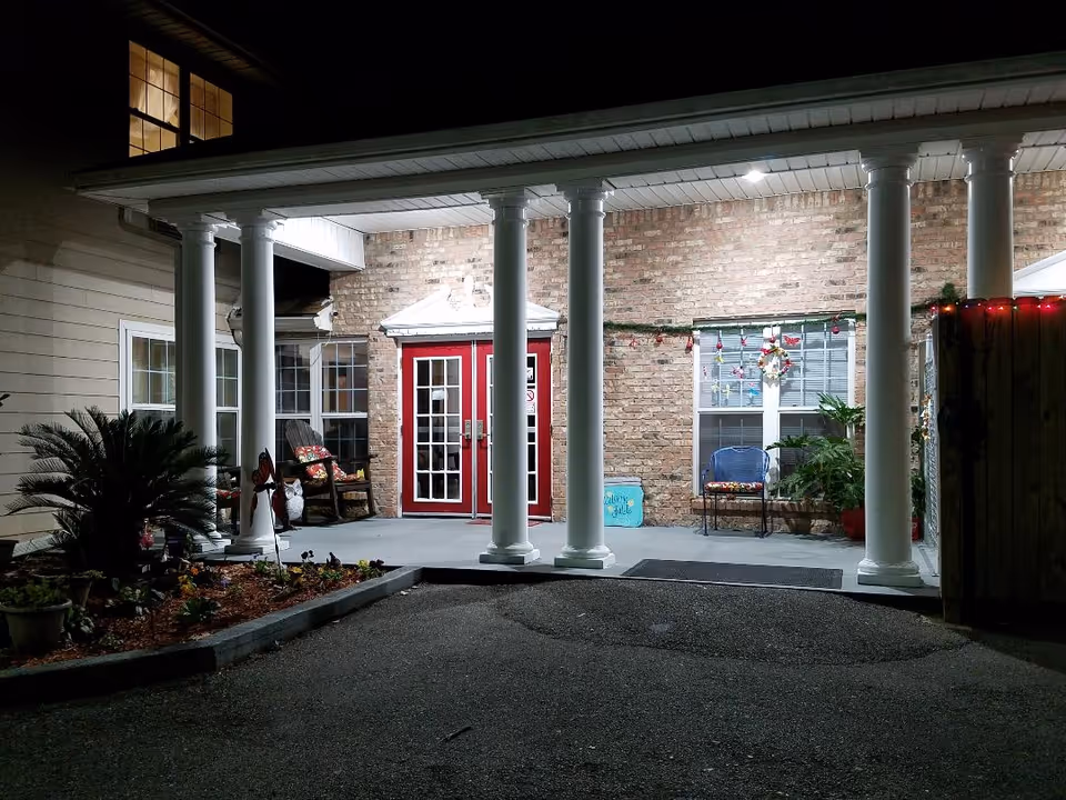 Night view of the entrance to a building with white columns supporting a covered porch. The entrance features double red doors with glass panels, a brick wall, and windows on either side. There are chairs with cushions and potted plants on the porch, along with some decorative items and a small garden bed in front.