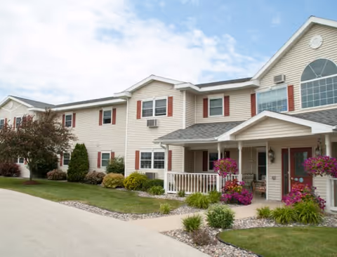 Front exterior of a two-story beige senior living building with red shutters, a covered entrance porch, and a landscaped lawn with flowering plants.
