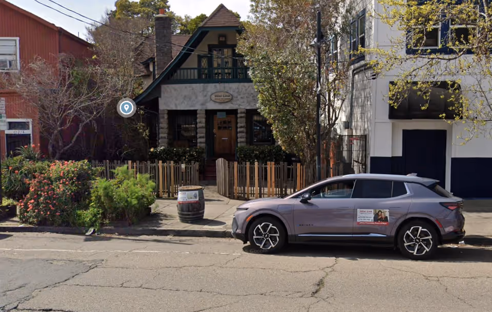 A small houselike building with a wooden fence and a parked SUV on the street.