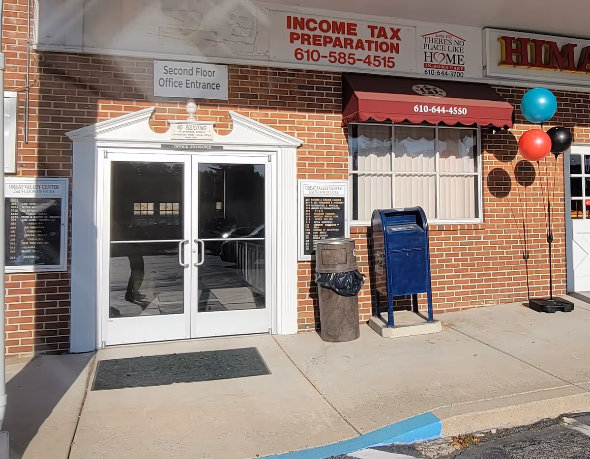 Brick storefront exterior with double glass doors, signs, a blue mailbox, and balloons.
