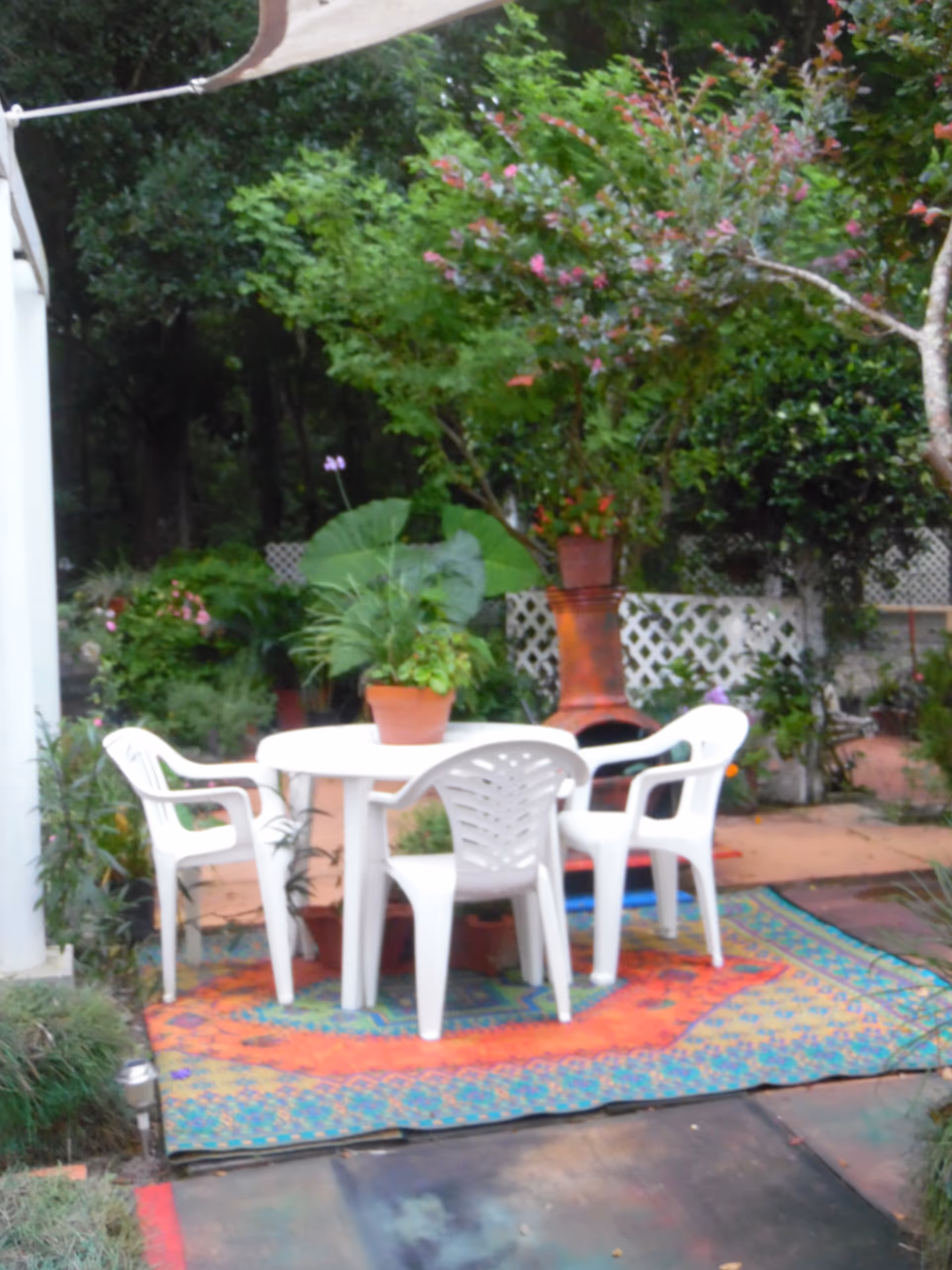 Outdoor patio with white plastic table and chairs, potted plants, and a colorful rug.