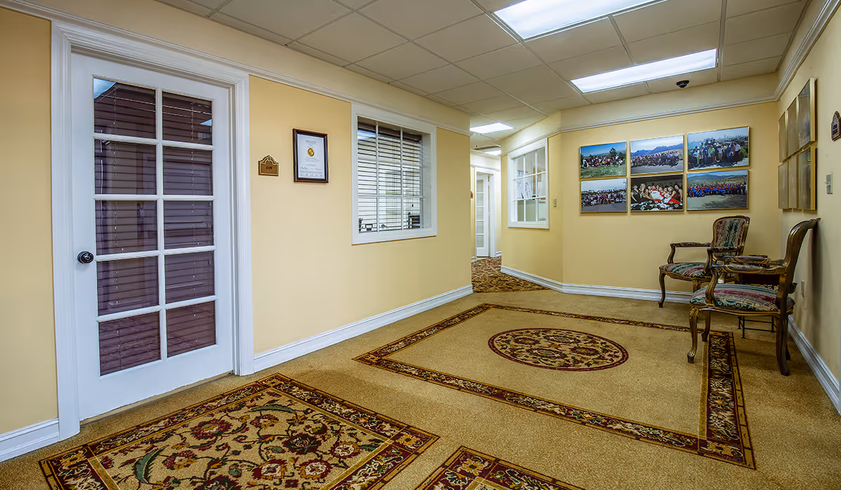 Yellow-walled hallway interior with framed photos, carpet, and wicker chairs along the wall.