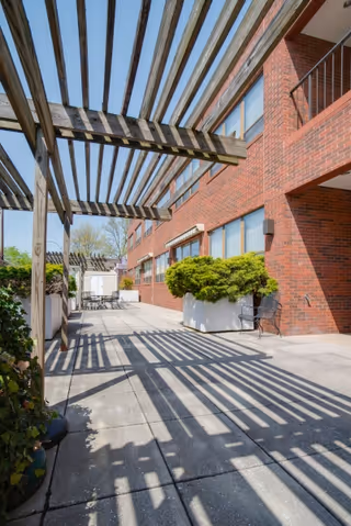 Brick courtyard with a wooden pergola casting striped shadows over a paved patio lined with planters and seating.
