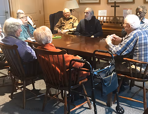 A group of elderly people sitting around a wooden table in a room, engaged in conversation. The room has wooden chairs, a lamp, framed pictures on the wall, and a walker is visible next to one of the chairs.