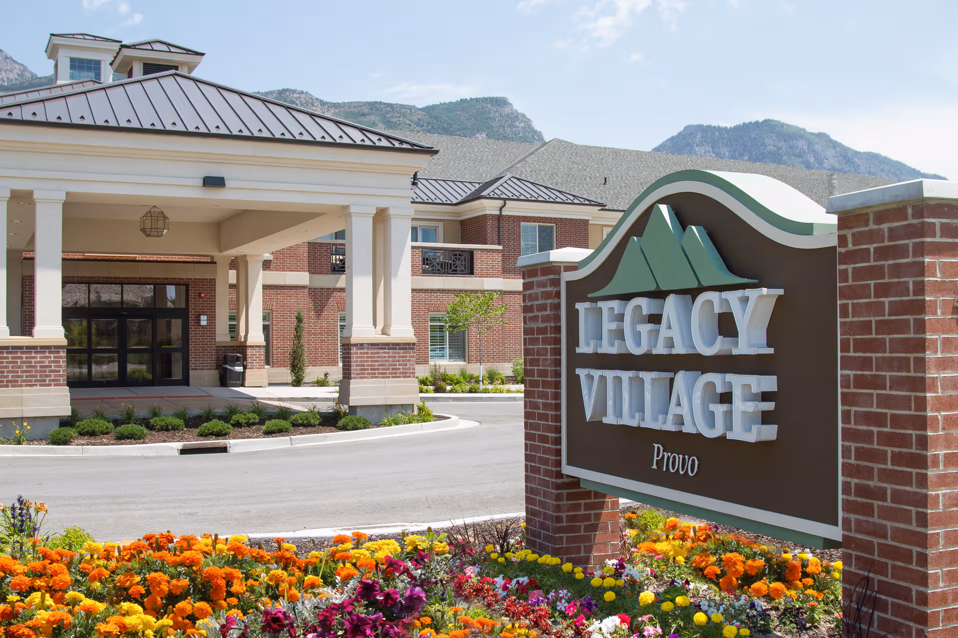 Front exterior of Legacy Village of Provo with a brick entrance, sign, and colorful flower beds.
